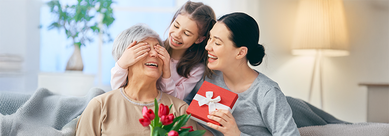 A trio of grandma, mother and daughter are smiling, the younger girl is covering her nan's eyes, the mother is holding a red-packaged gift with a white bow, and the grandmother is holding flowers.